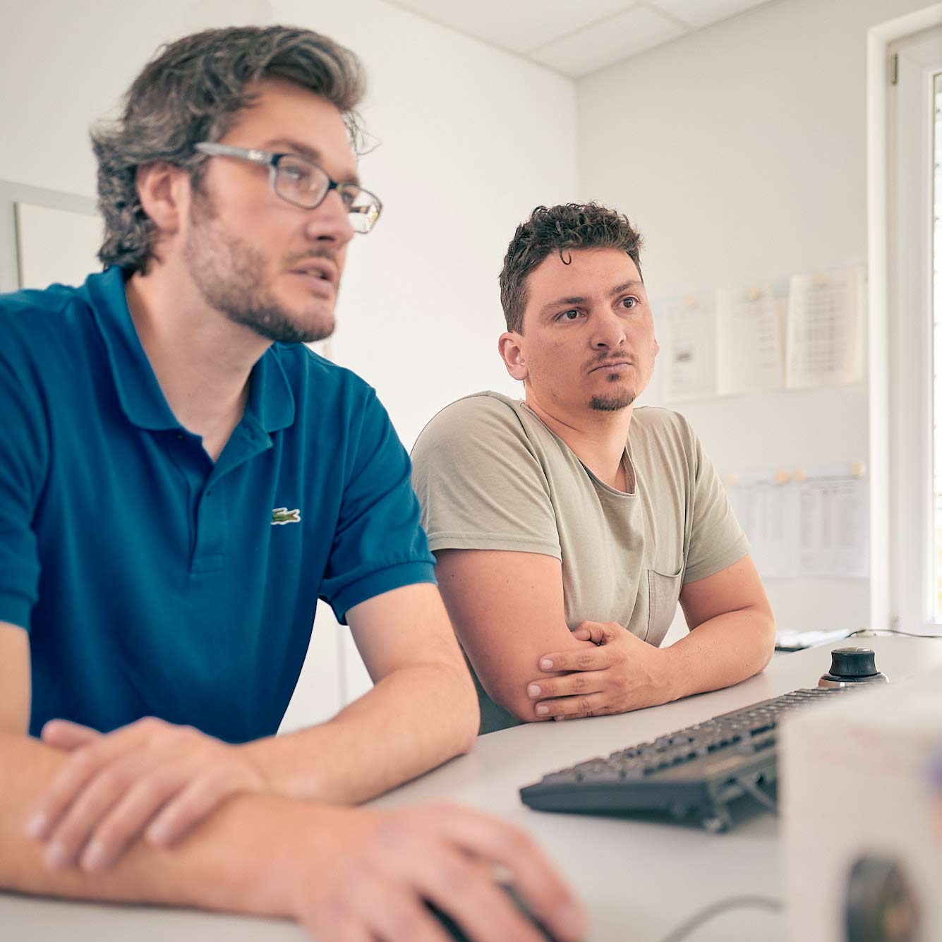 Zwei Mitarbeiter der M Consult GmbH tauschen sich aus. Sie stehen an einem Schreibtisch und betrachten einen Monitor.